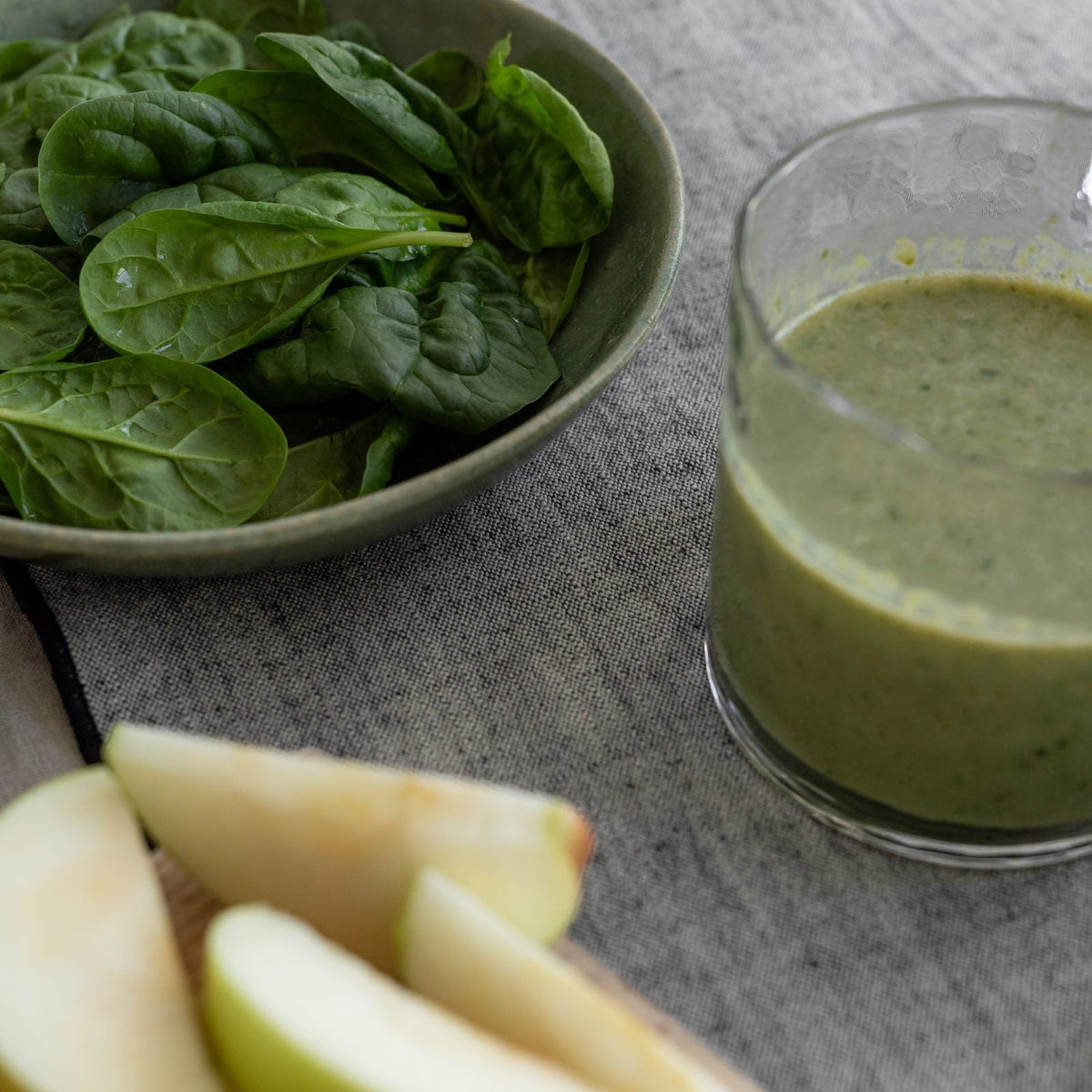 A bowl of fresh spinach beside a green smoothie, sliced apple and lime