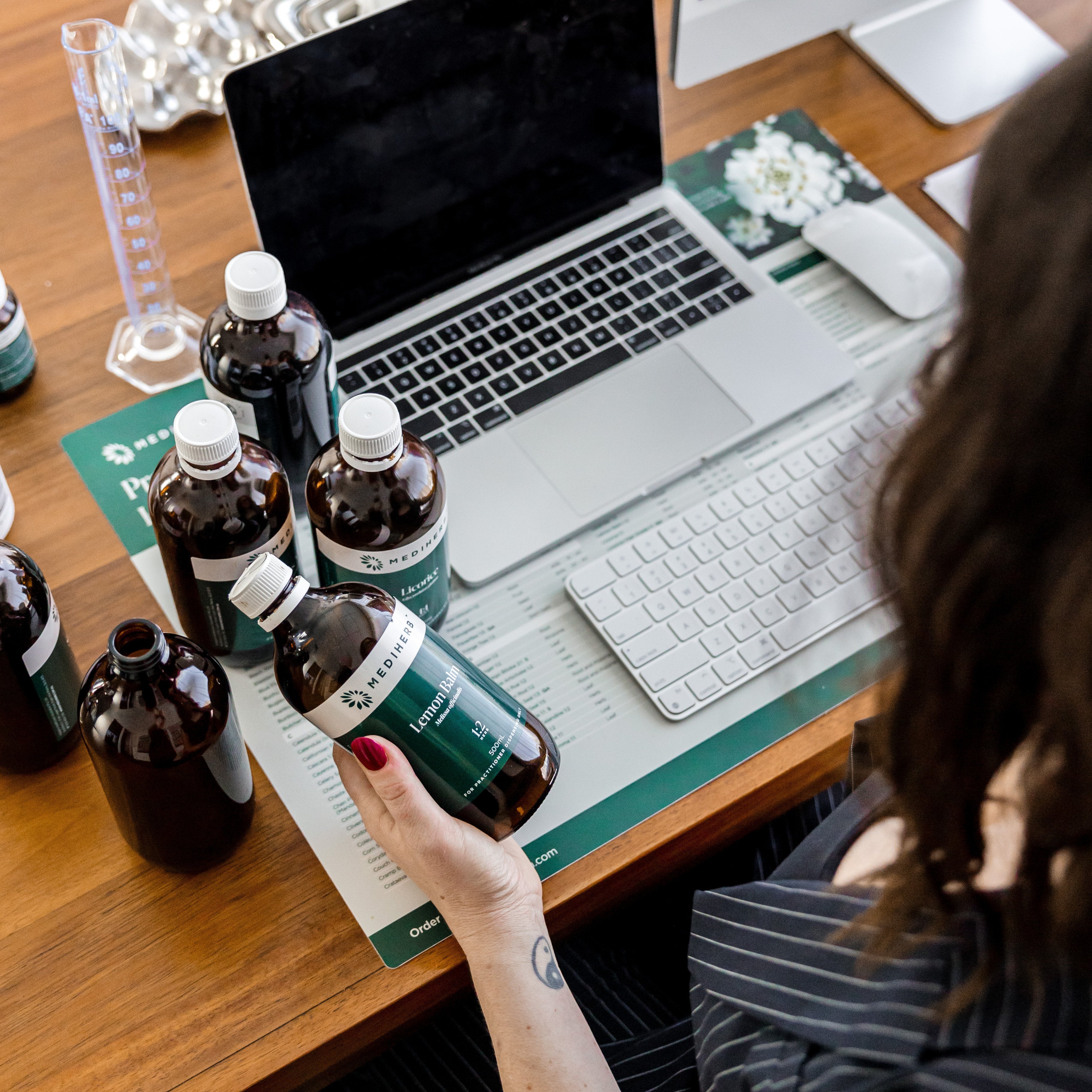 A natural health practitioner holds a herbal medicine bottle while working at a desk with custom formulations, supplements, and a laptop in a modern apothecary.