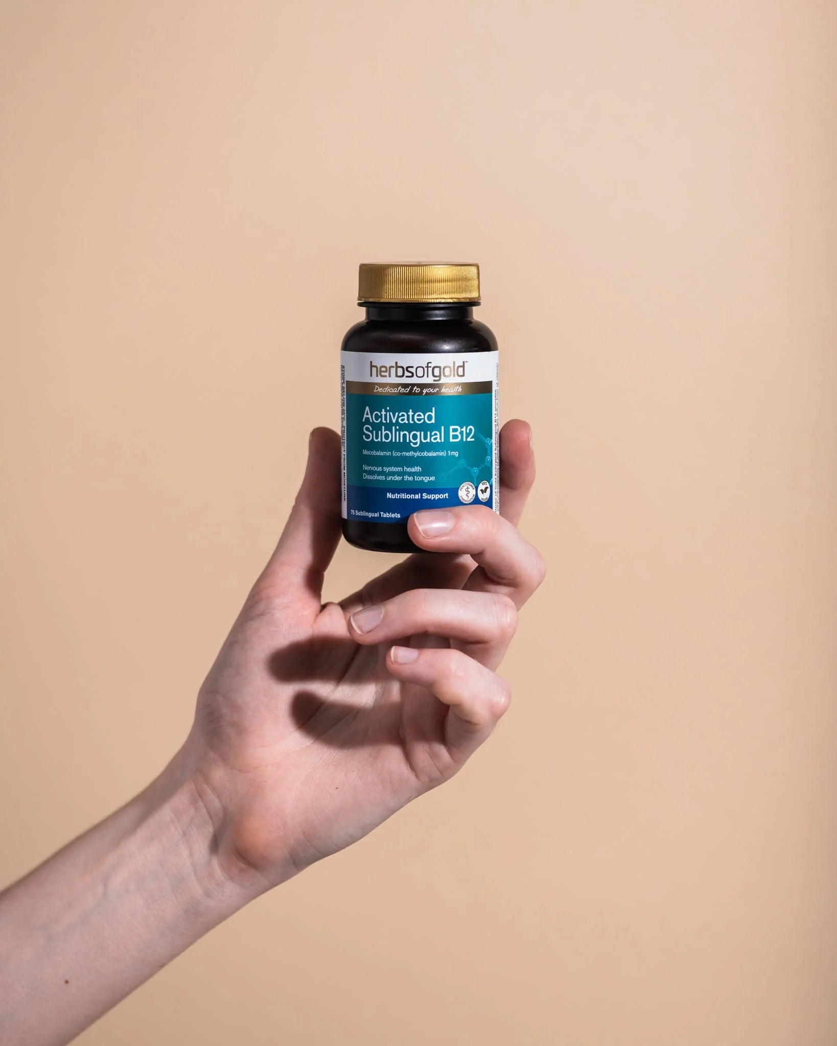 Person holding Herbs of Gold Activated Sublingual B12 bottle upright against a soft beige background.