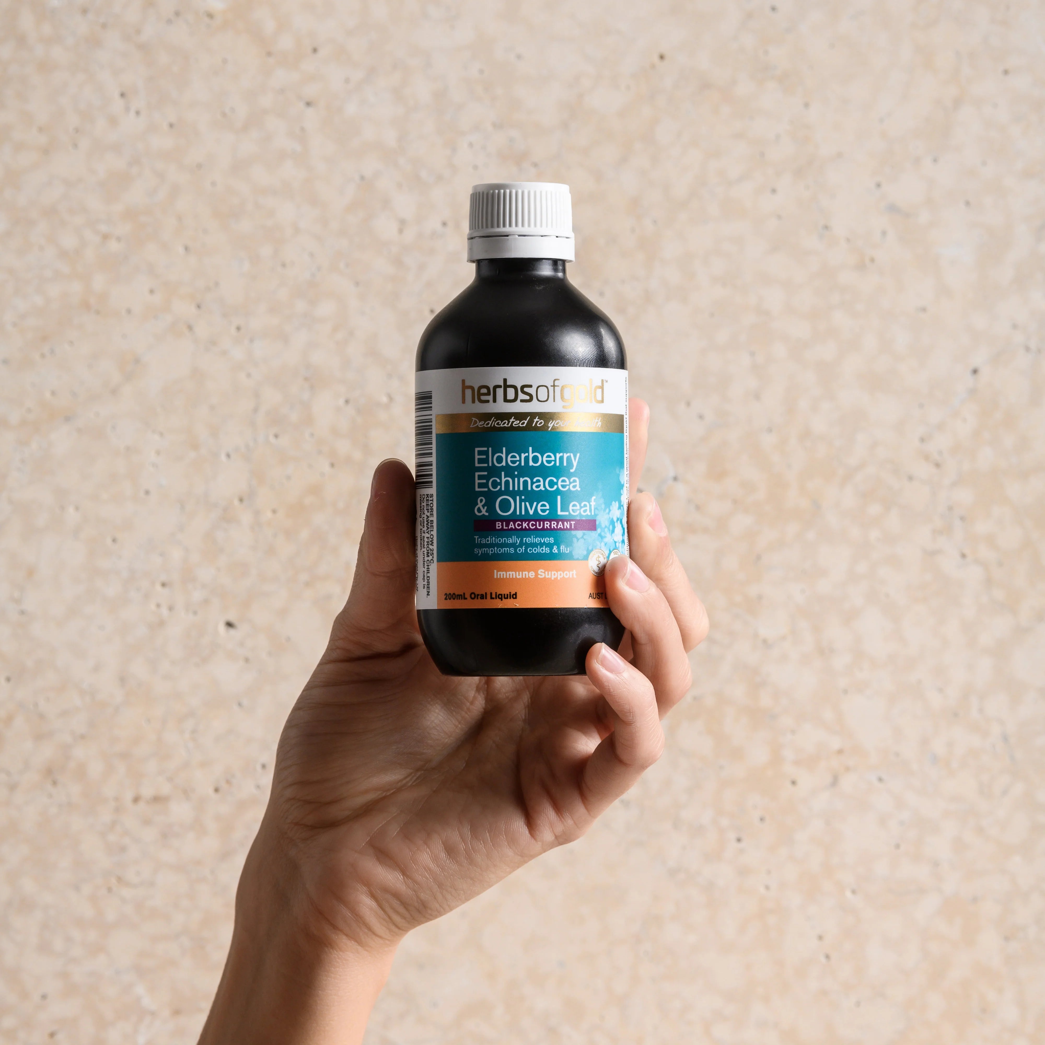 Hand holding a bottle of Herbs of Gold Elderberry Echinacea & Olive Leaf supplement against a beige background