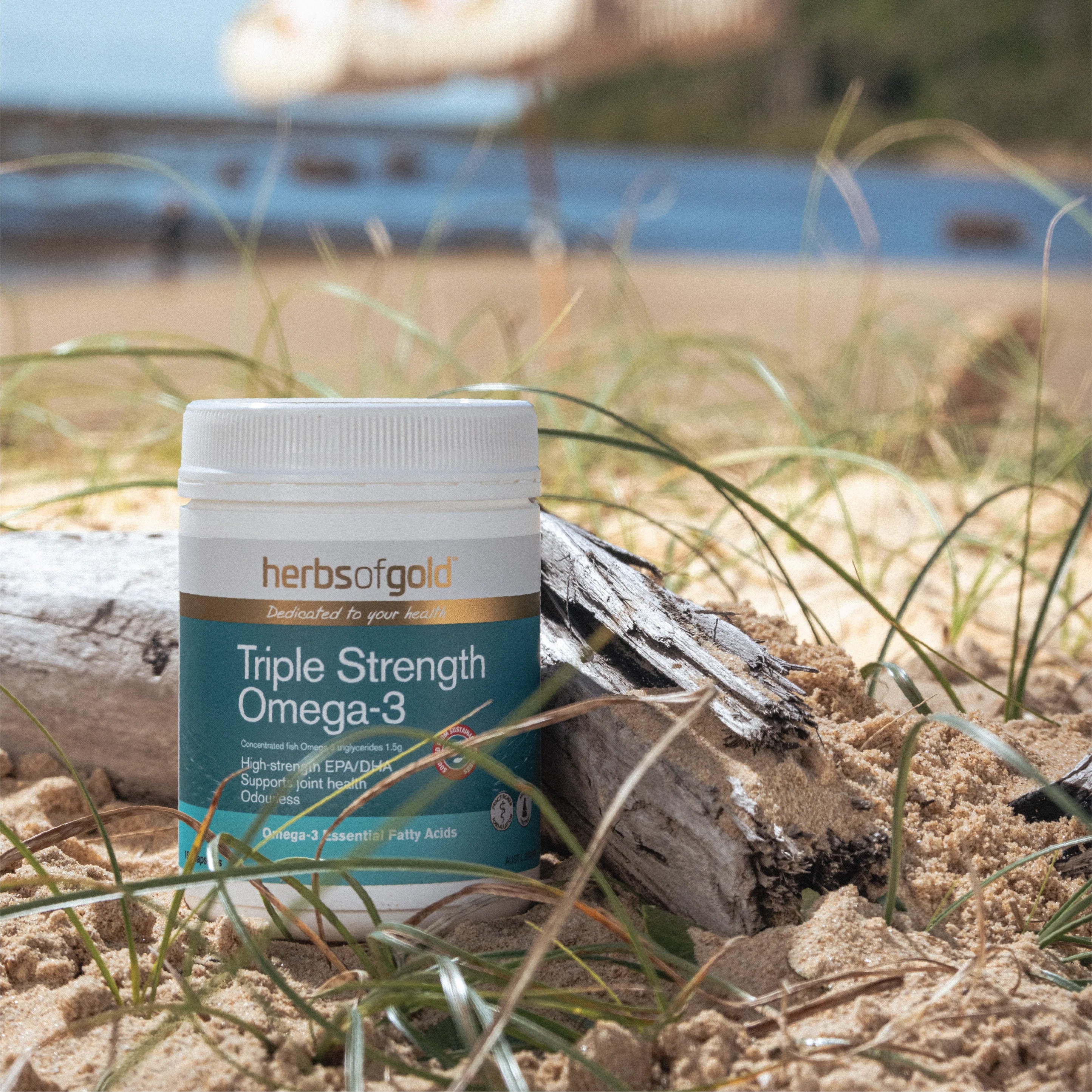 Herbs of Gold Triple Strength Omega-3 supplement bottle on a beach with grass and water in the background