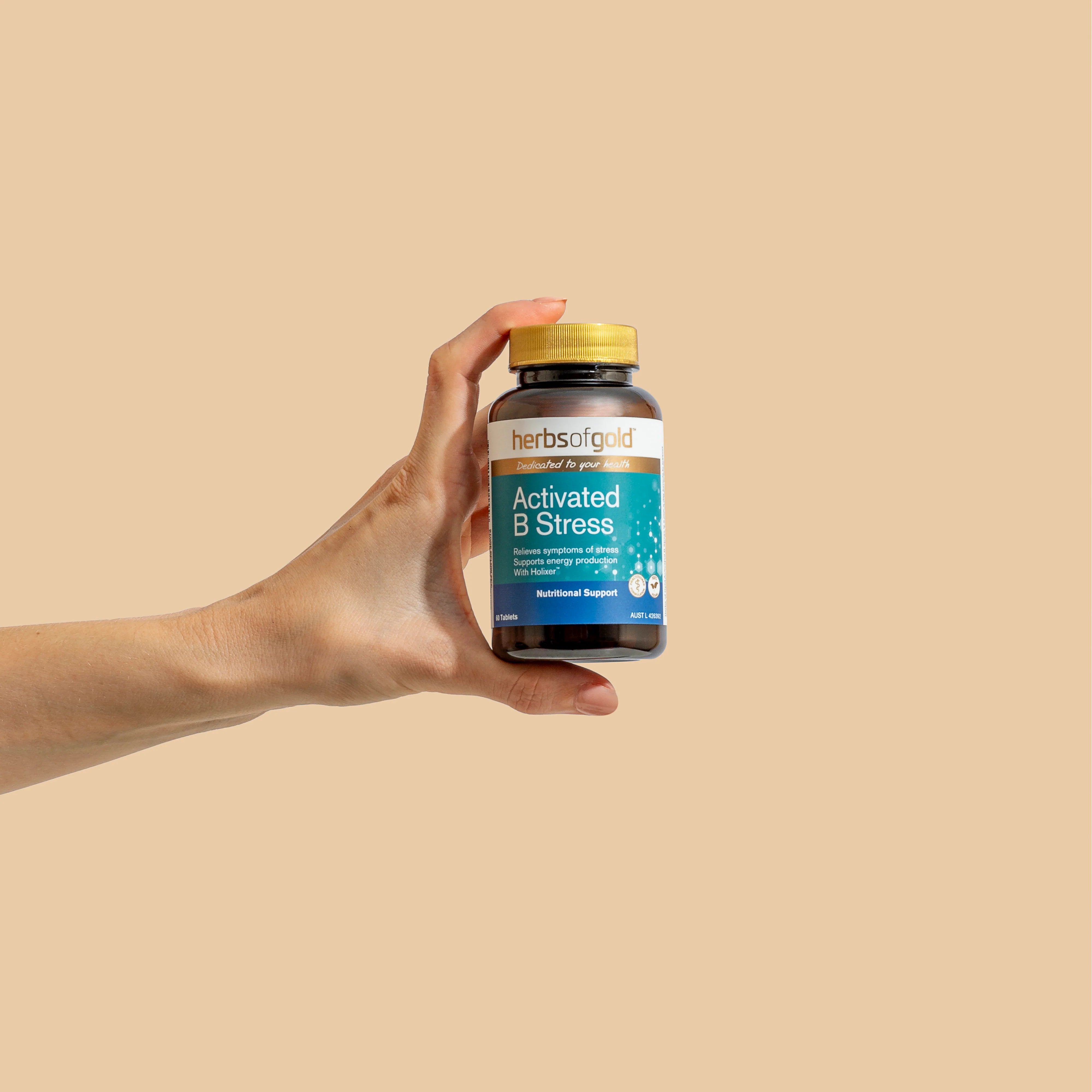 Hand holding a Herbs of Gold Activated B Stress bottle upright against a beige background.