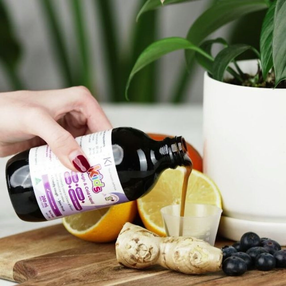 Hand pouring a liquid from a bottle labeled 'KIDS' into a small cup with fruits and a plant in the background.