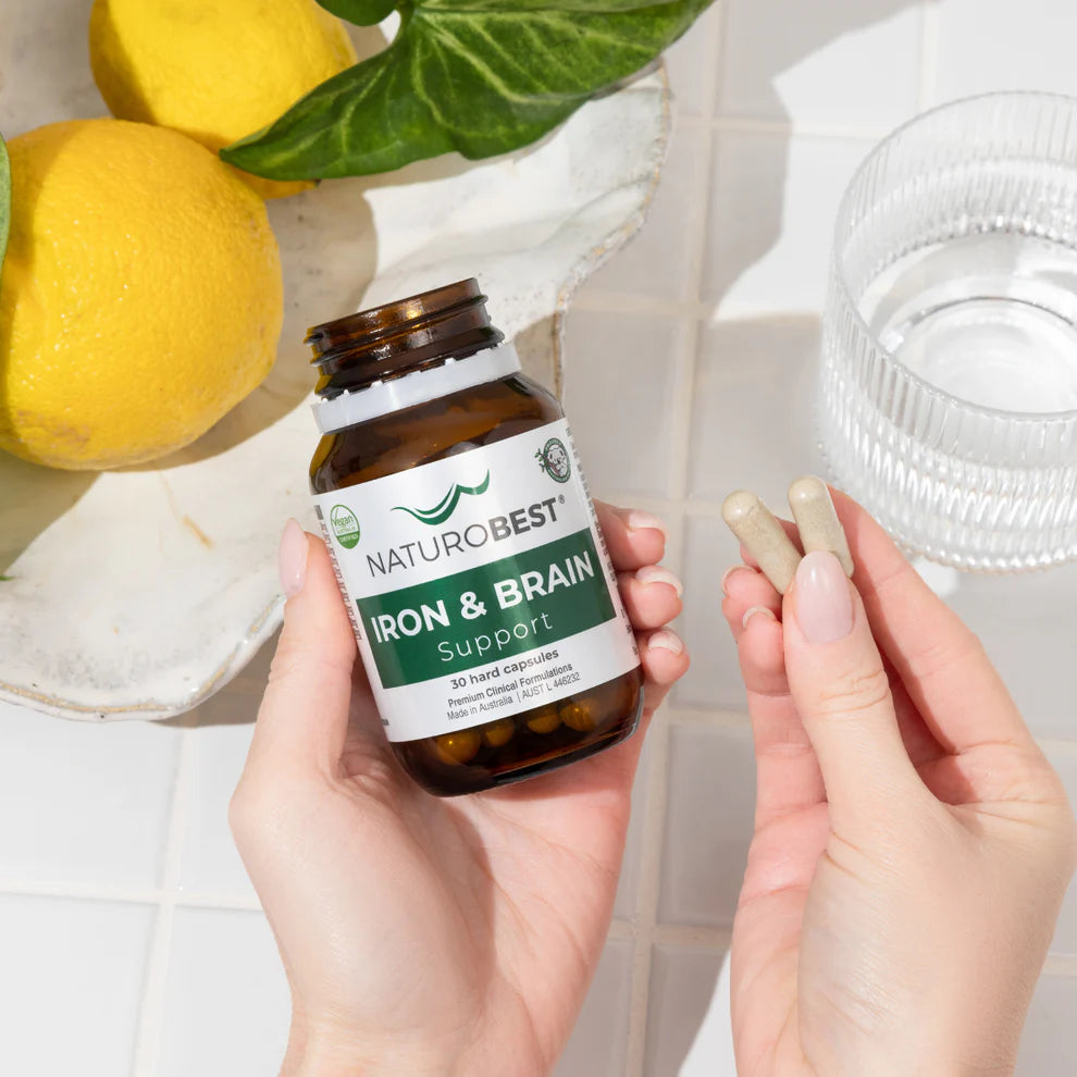 Person holding a bottle of NaturoBest Iron & Brain Support next to fresh lemons and a glass of water, highlighting its natural, wellness-focused presentation.