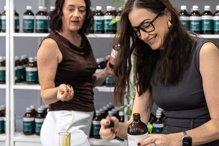 Two women in a store setting with shelves of bottles at Nourishing Apothecary.