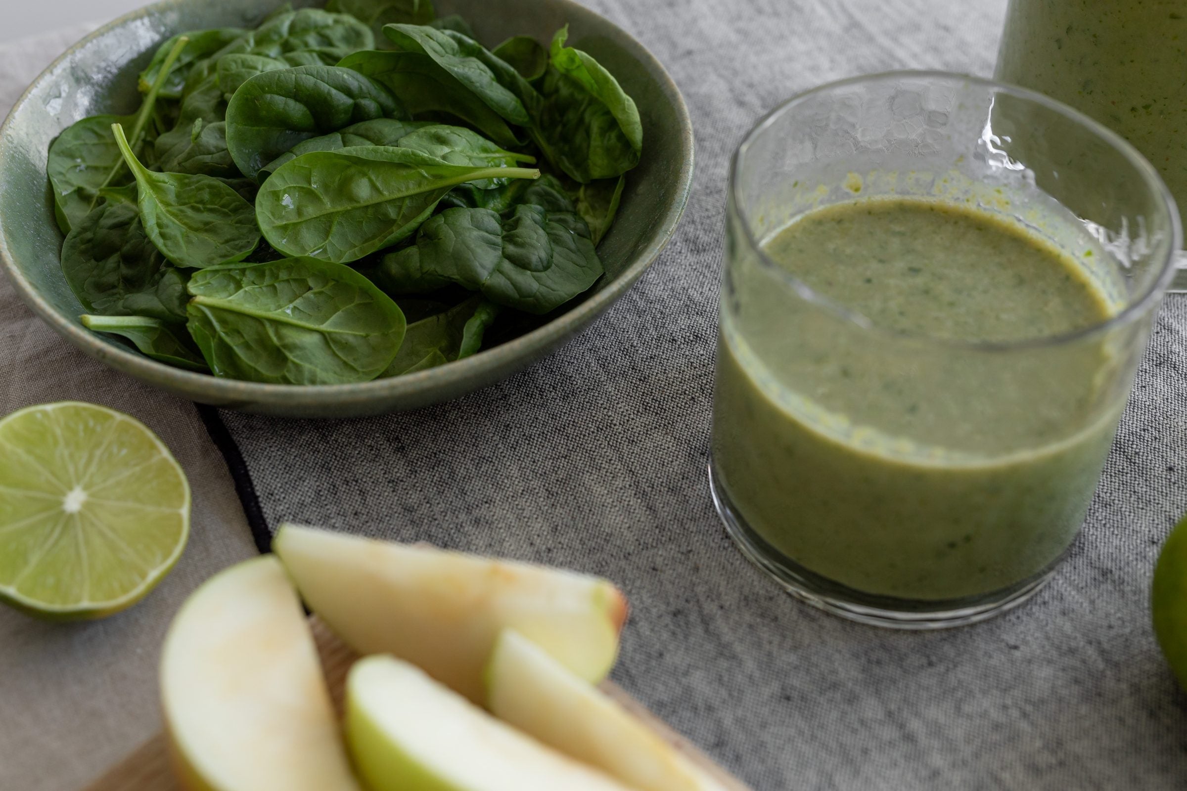 A bowl of fresh spinach beside a green smoothie, sliced apple and lime