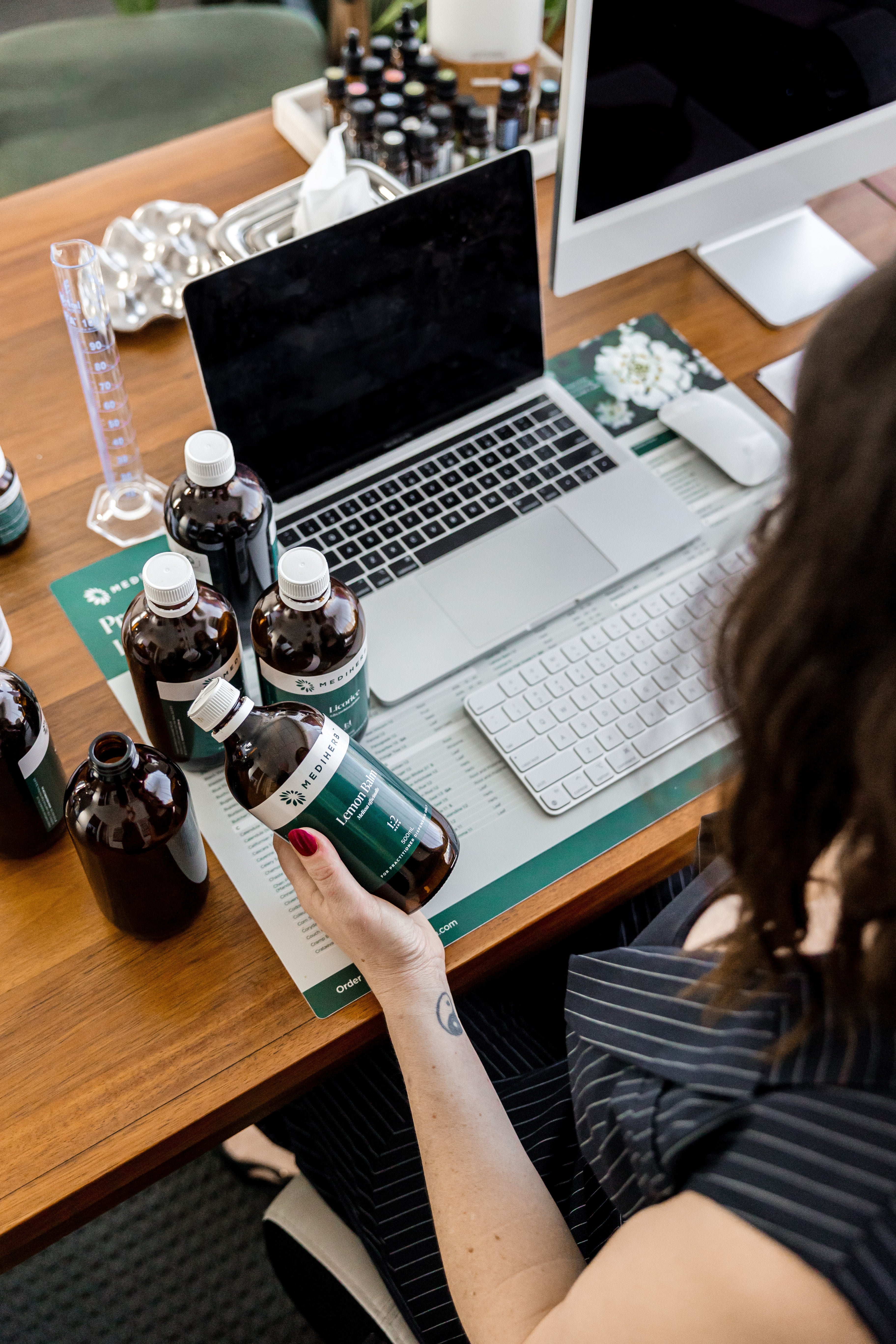 A natural health practitioner holds a herbal medicine bottle while working at a desk with custom formulations, supplements, and a laptop in a modern apothecary.