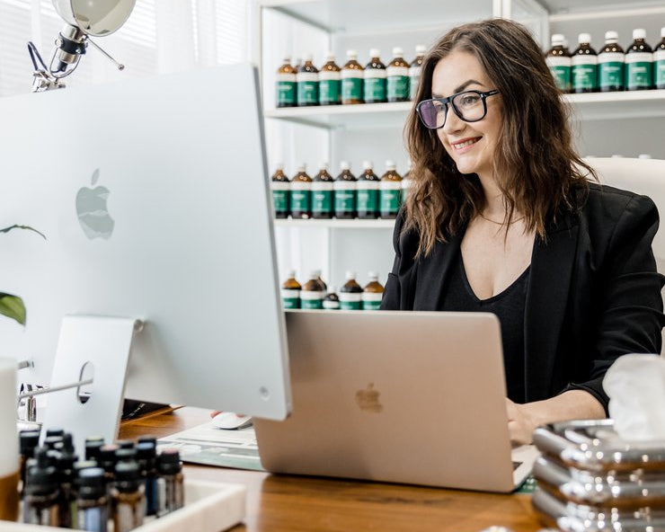 Woman sitting at a desk with a laptop, surrounded by bottles on shelves.